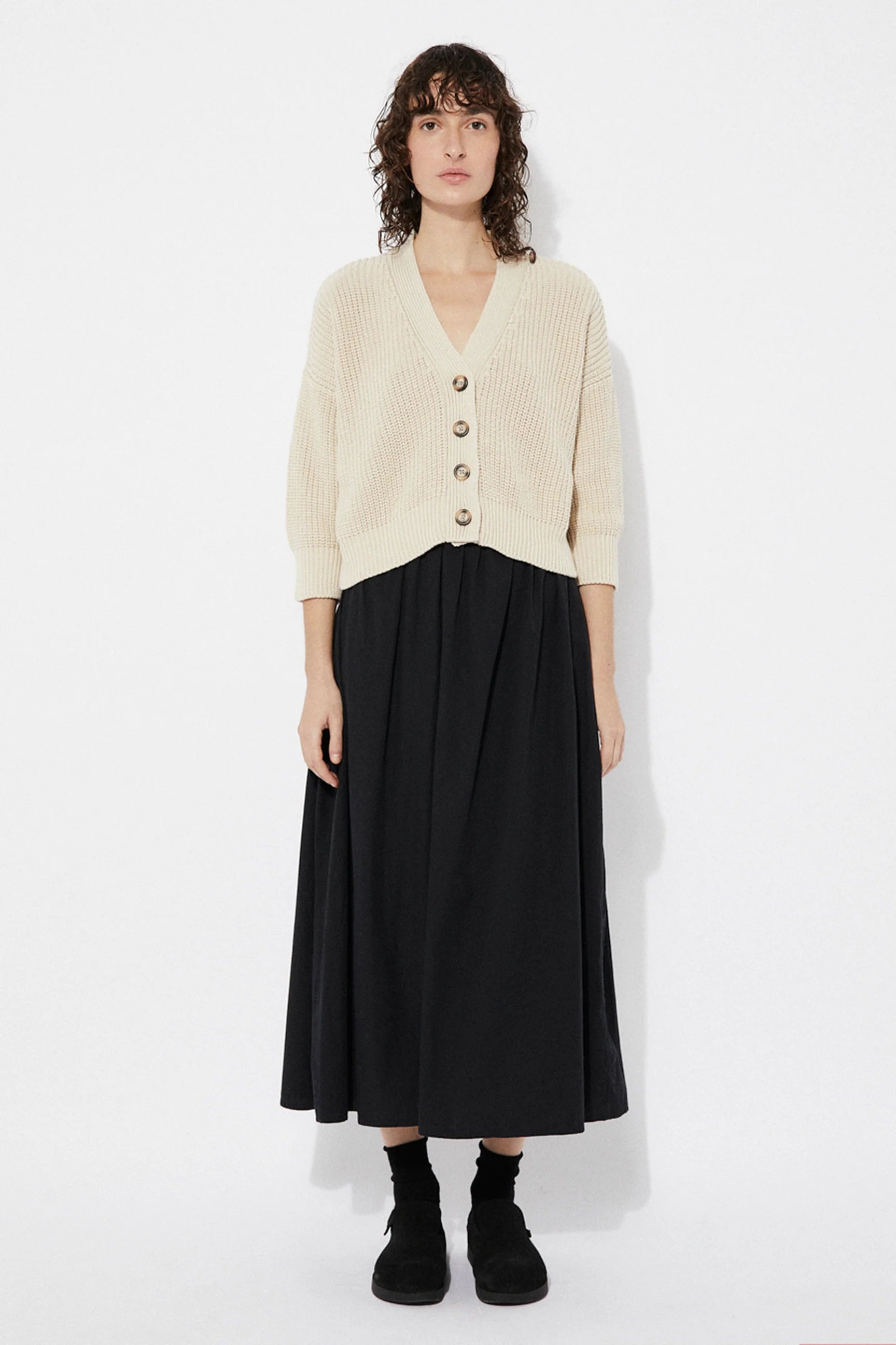 Woman wearing a beige Sclater knitted cardigan by Rita Row with buttons with a black skirt on a white background