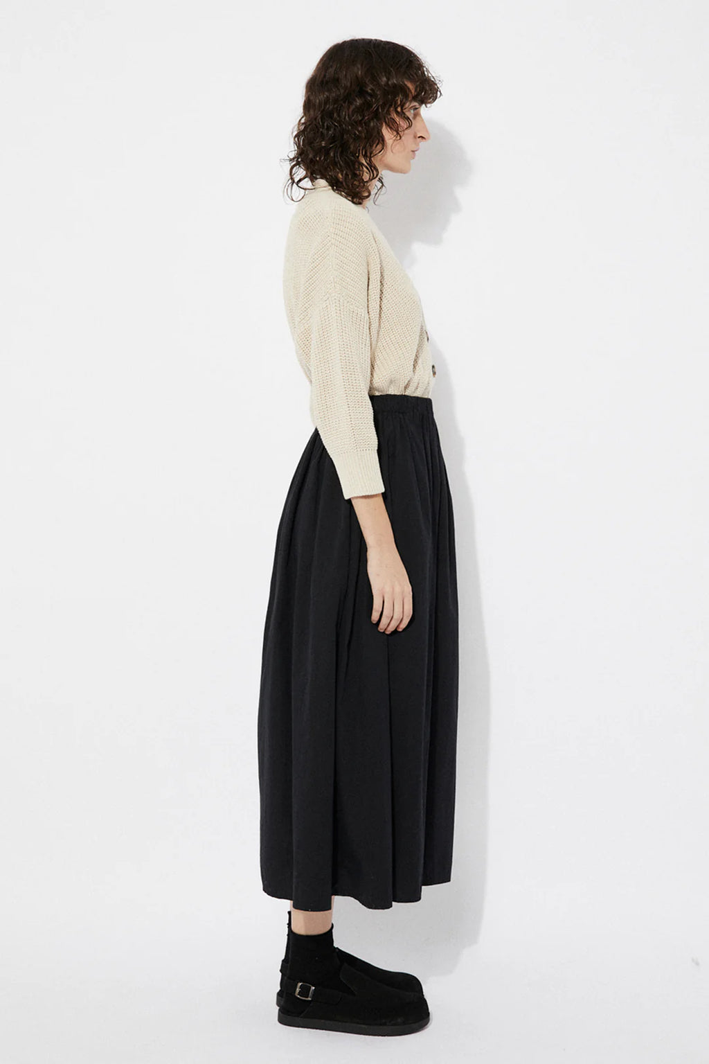 Woman wearing a beige Sclater knitted cardigan by Rita Row with buttons with a black skirt on a white background
