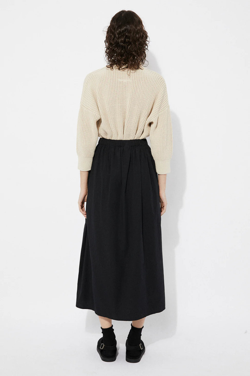 Woman wearing a beige Sclater knitted cardigan by Rita Row with buttons with a black skirt on a white background