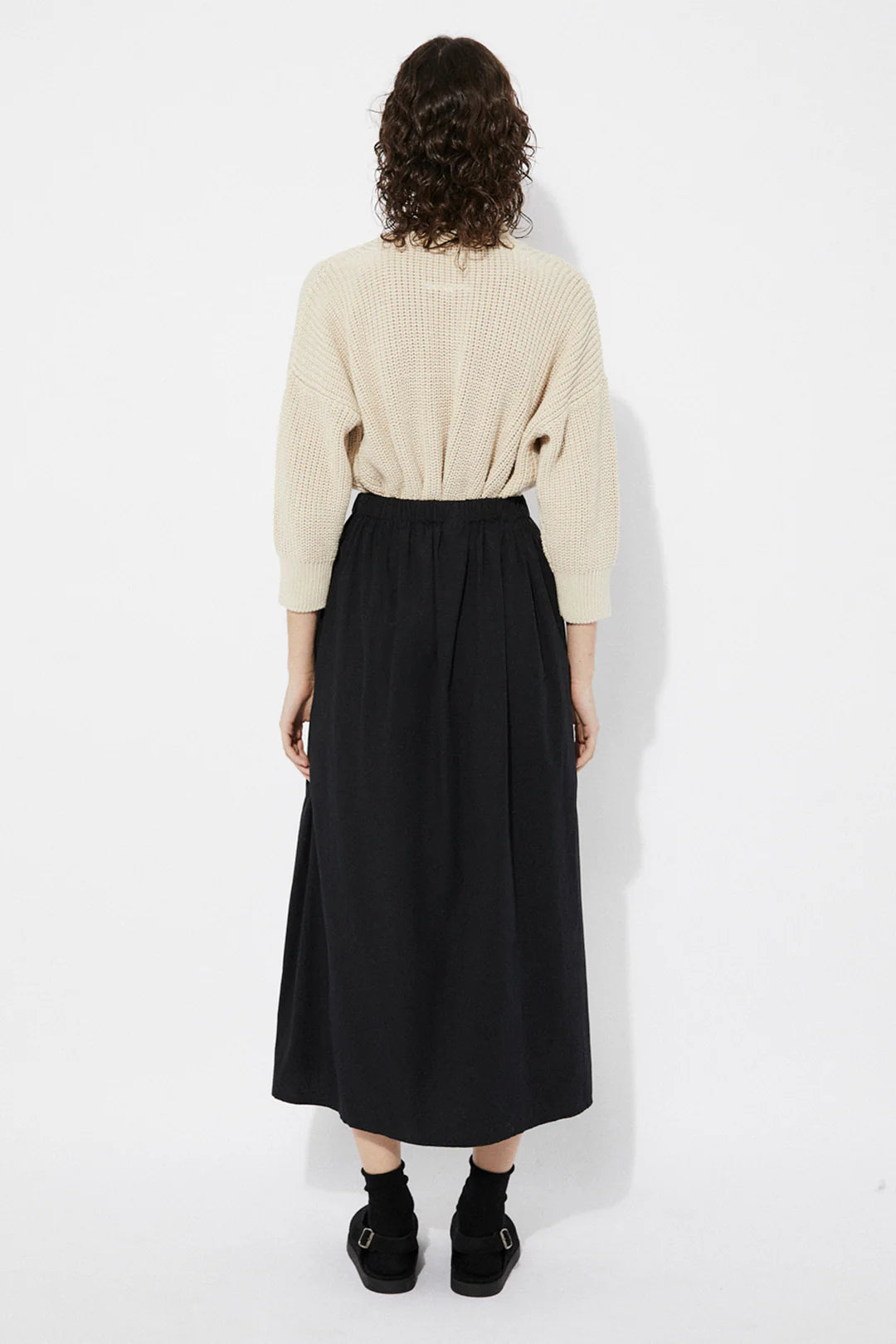 Woman wearing a beige Sclater knitted cardigan by Rita Row with buttons with a black skirt on a white background