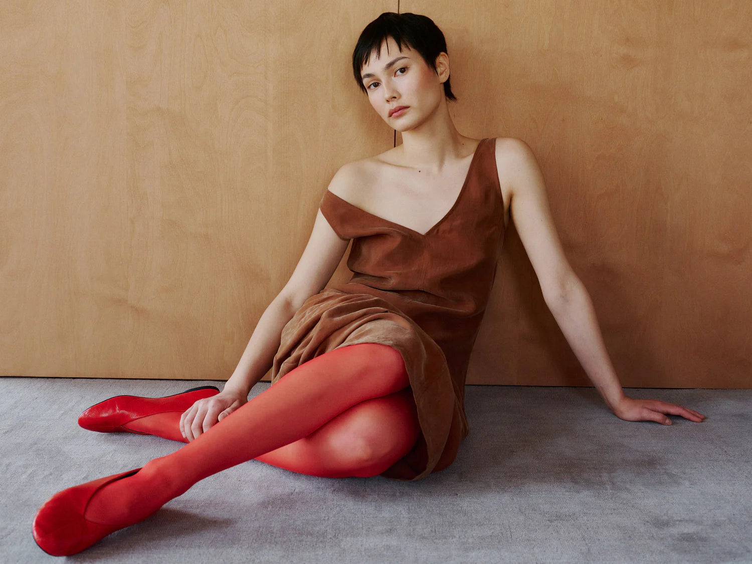 Woman in a brown dress and red tights sitting on the floor against a beige wall.