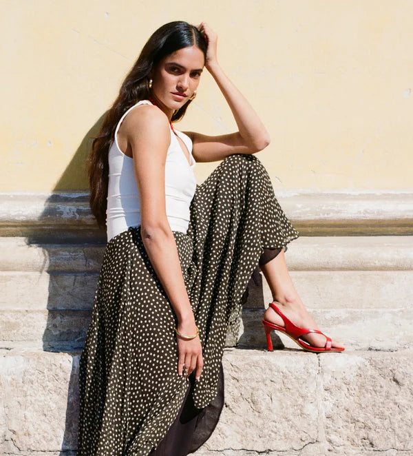 A woman sits on stone steps against a pale yellow wall, wearing a white sleeveless top and a long dark skirt with small light polka dots, paired with red high-heeled sandals.