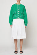 Woman wearing a green sweater jacket with lilac stitching by Atelier Delphine and a white skirt on a plain background