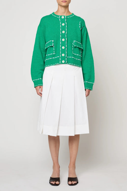 Woman wearing a green sweater jacket with lilac stitching by Atelier Delphine and a white skirt on a plain background