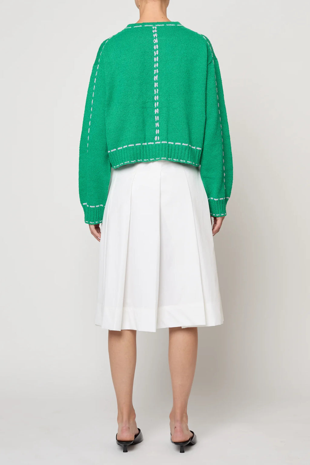 Woman wearing a green sweater jacket with lilac stitching by Atelier Delphine and a white skirt on a plain background