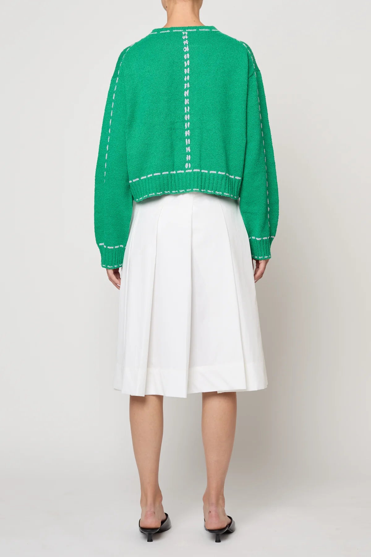 Woman wearing a green sweater jacket with lilac stitching by Atelier Delphine and a white skirt on a plain background