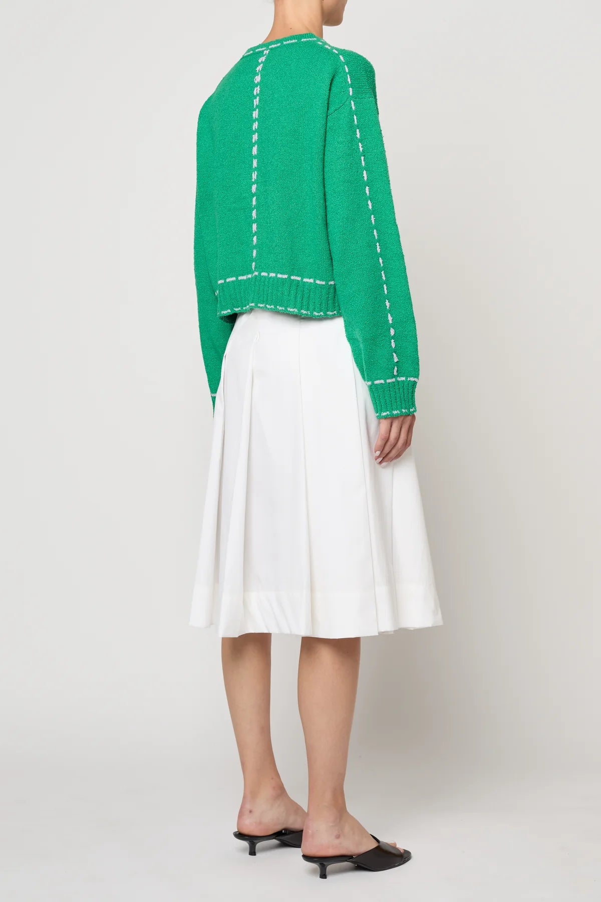 Woman wearing a green sweater jacket with lilac stitching by Atelier Delphine and a white skirt on a plain background
