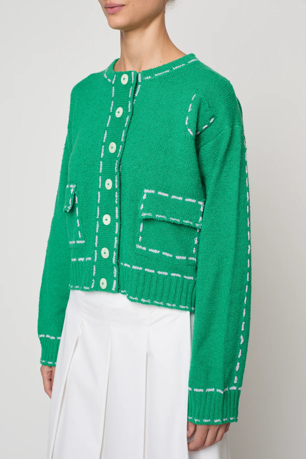 Woman wearing a green sweater jacket with lilac stitching by Atelier Delphine and a white skirt on a plain background