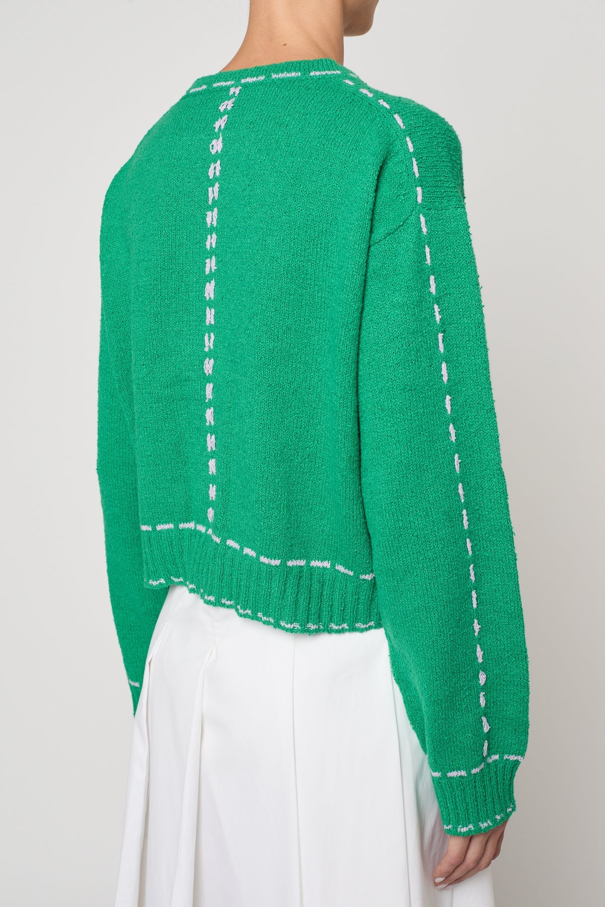 Woman wearing a green sweater jacket with lilac stitching by Atelier Delphine and a white skirt on a plain background