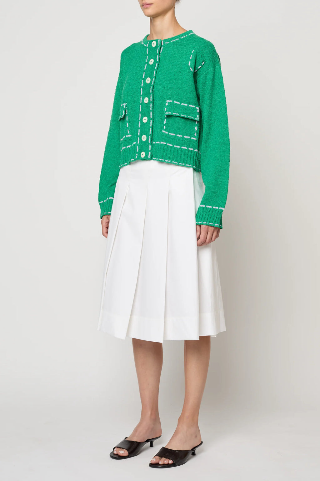 Woman wearing a green sweater jacket with lilac stitching by Atelier Delphine and a white skirt on a plain background