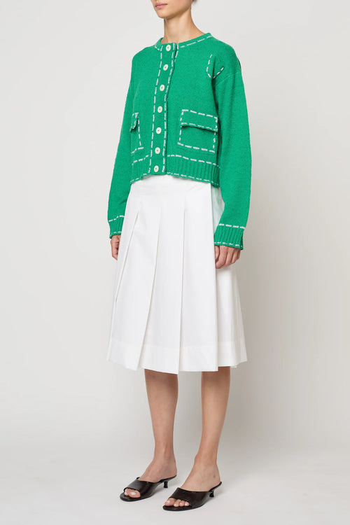 Woman wearing a green sweater jacket with lilac stitching by Atelier Delphine and a white skirt on a plain background