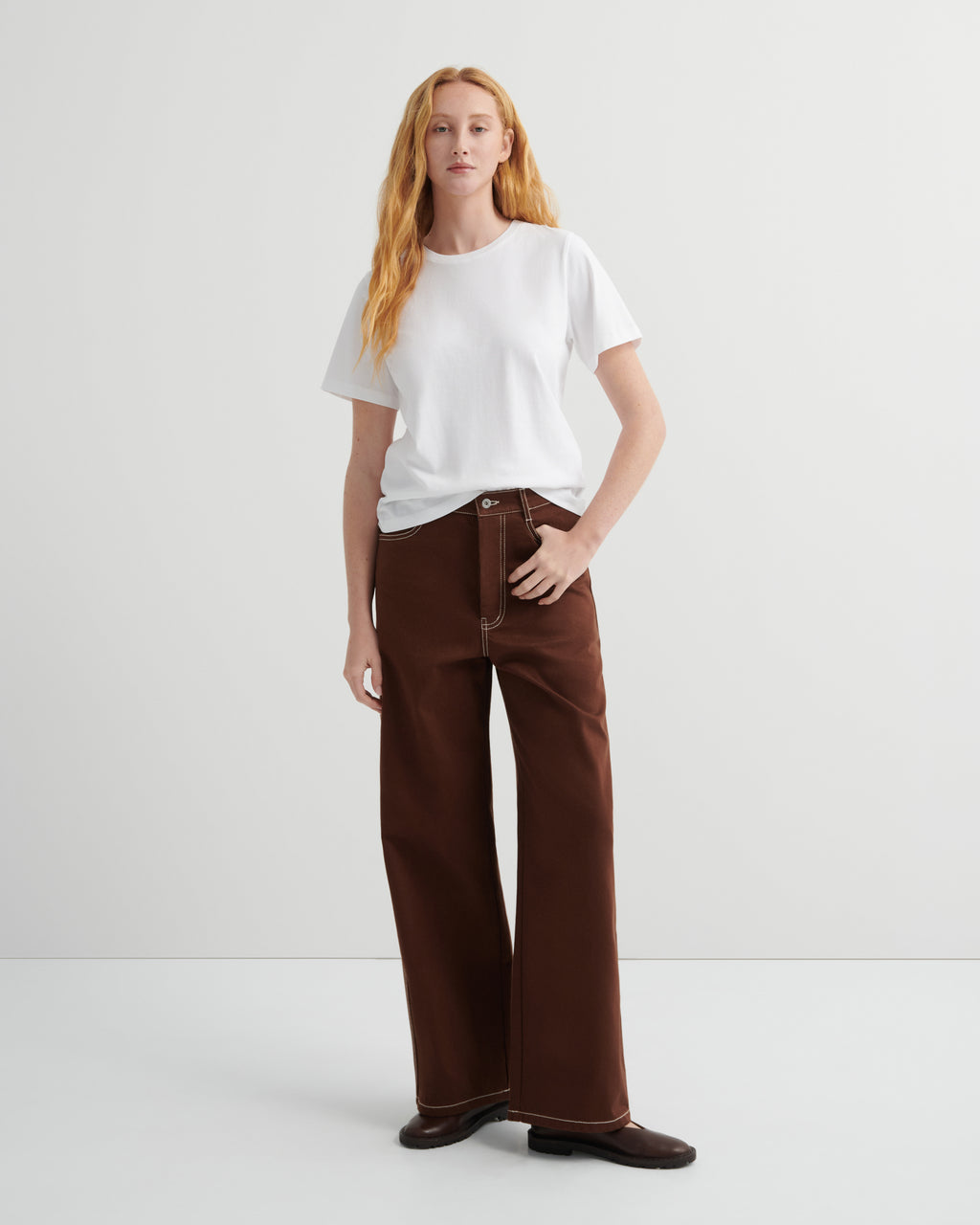 Woman wearing brown craft jeans by kowtow with a white shirt on a white background.