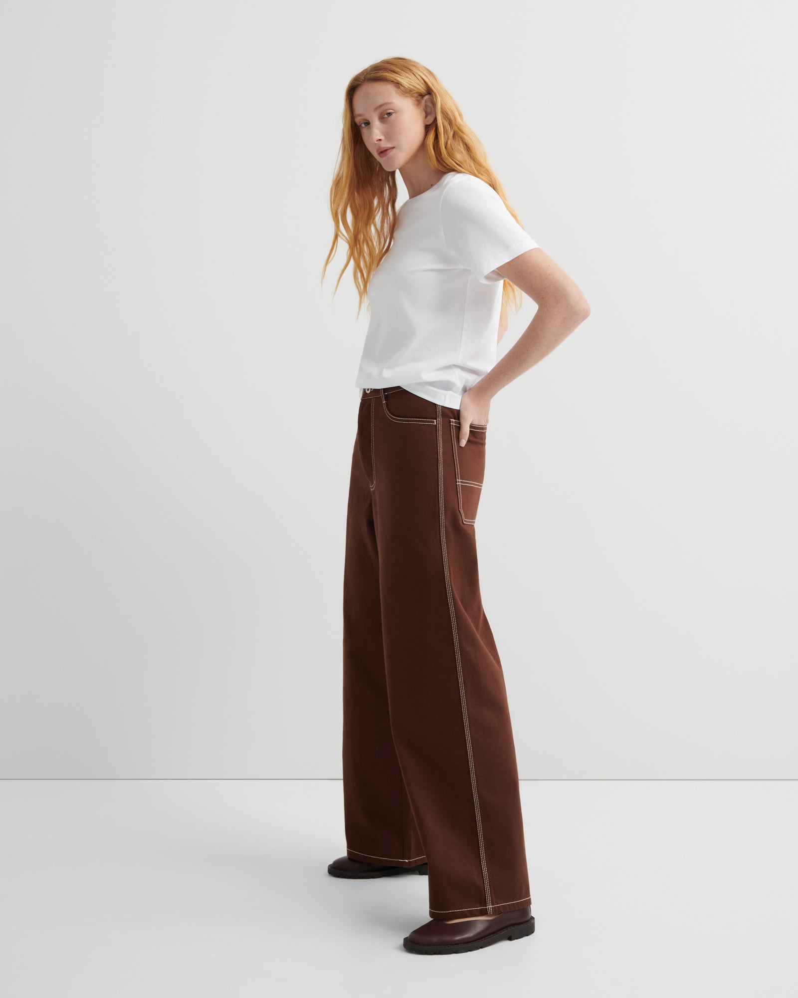 Woman wearing brown craft jeans by kowtow with a white shirt on a white background.