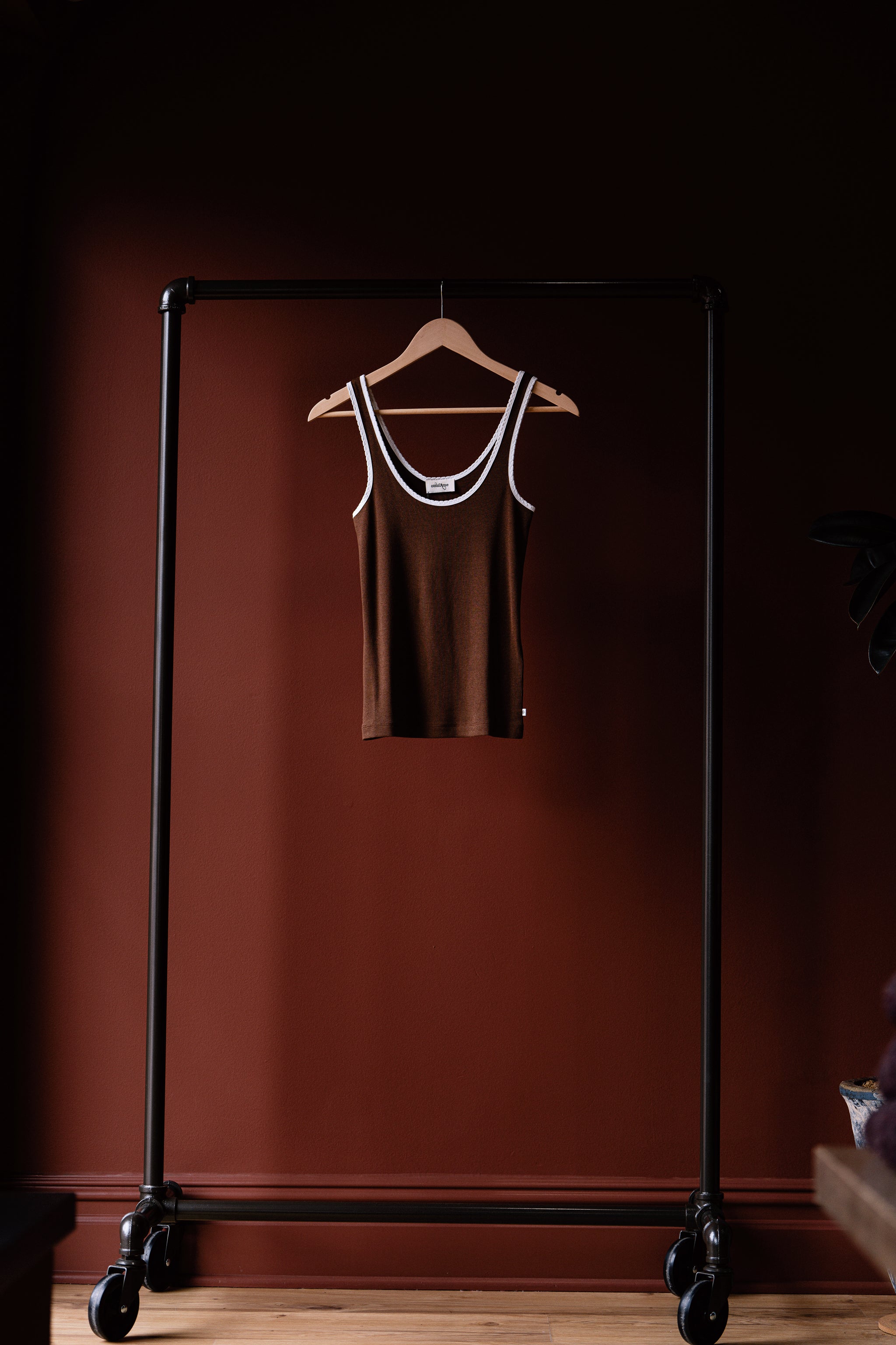 Brown tank top on a hanger attached to a black garment rack against a dark red wall.