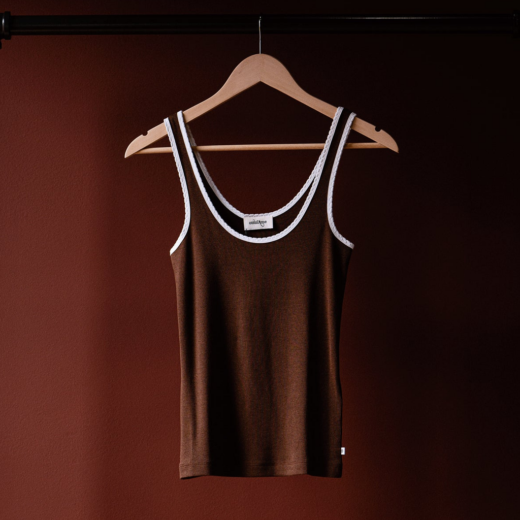 A brown ribbed tank top with white trim on the scoop neck and armholes, hanging on a wooden hanger against a dark reddish-brown background.