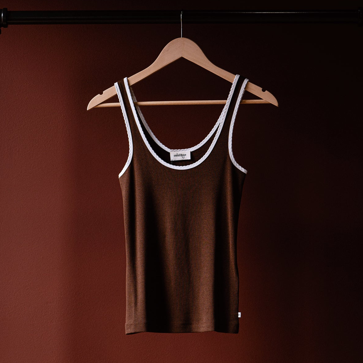 A brown ribbed tank top with white trim on the scoop neck and armholes, hanging on a wooden hanger against a dark reddish-brown background.