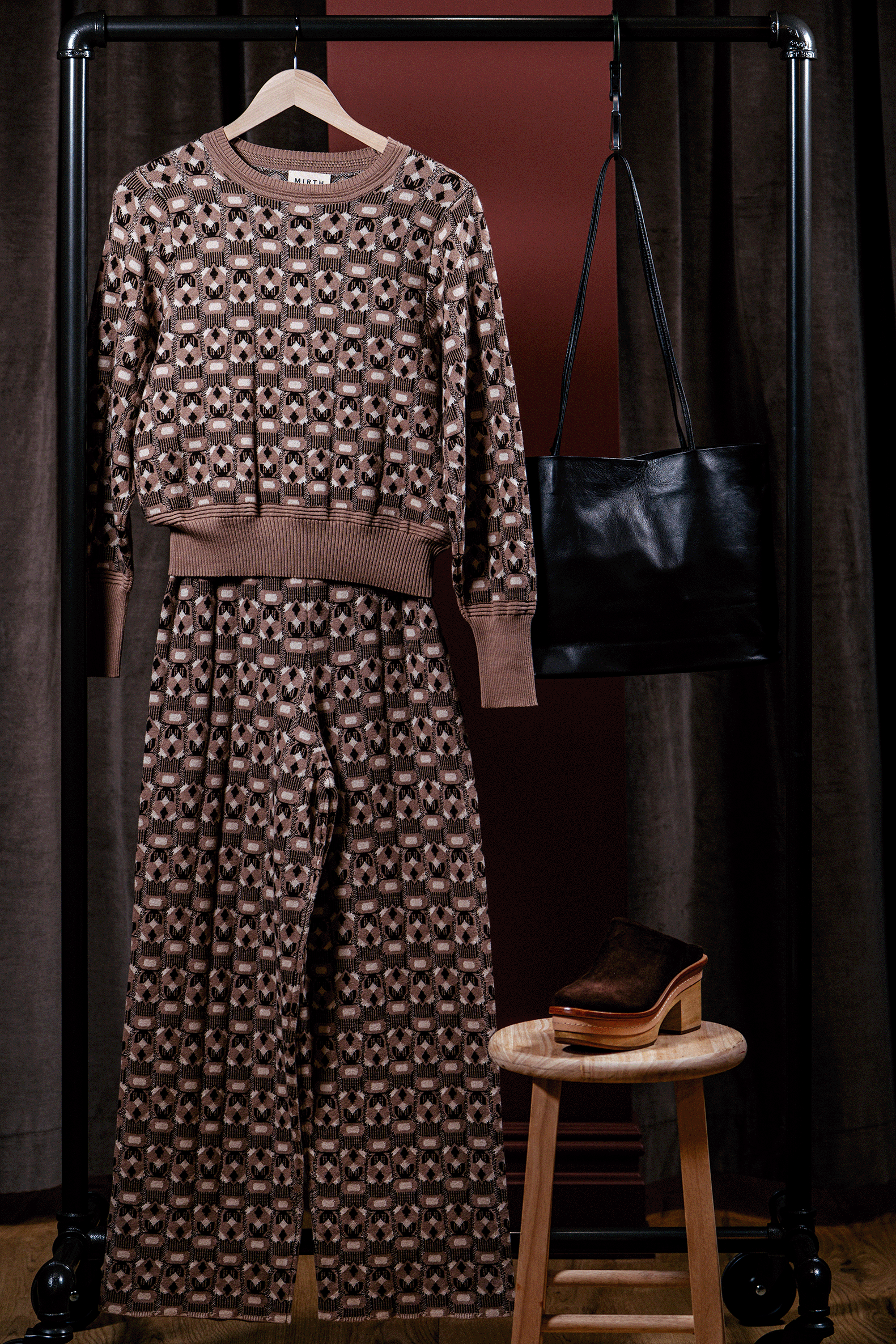 Patterned outfit with a sweater and pants on a rack, accompanied by a black handbag and wooden stool.