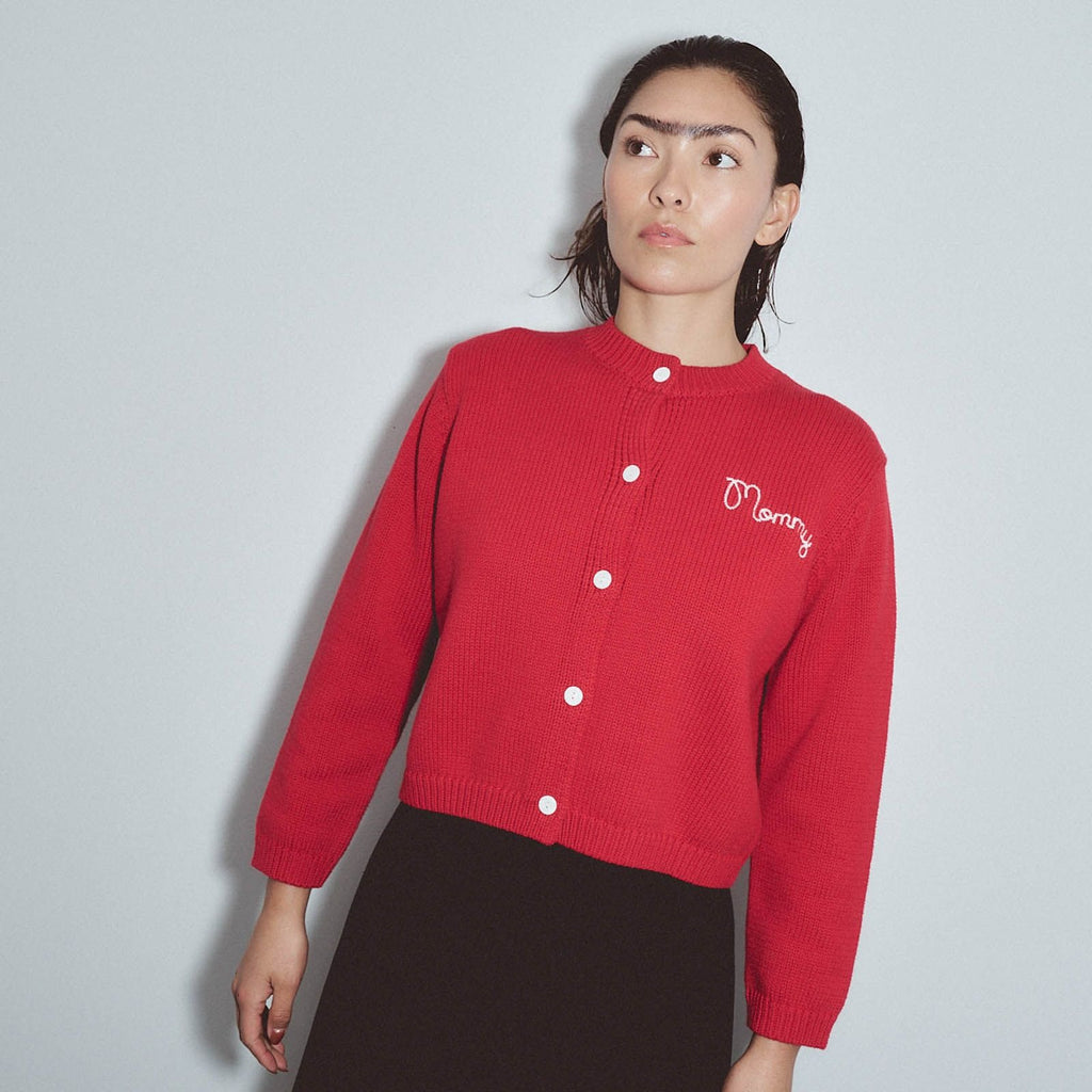 A woman wearing a red ribbed knit cardigan with white buttons and a white embroidered 'Merry' logo on the chest, paired with a dark brown skirt, against a light gray background. Mr. Rogers Mommy Cardigan by Fashion Brand Company.