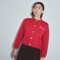 A woman wearing a red ribbed knit cardigan with white buttons and a white embroidered 'Merry' logo on the chest, paired with a dark brown skirt, against a light gray background. Mr. Rogers Mommy Cardigan by Fashion Brand Company.