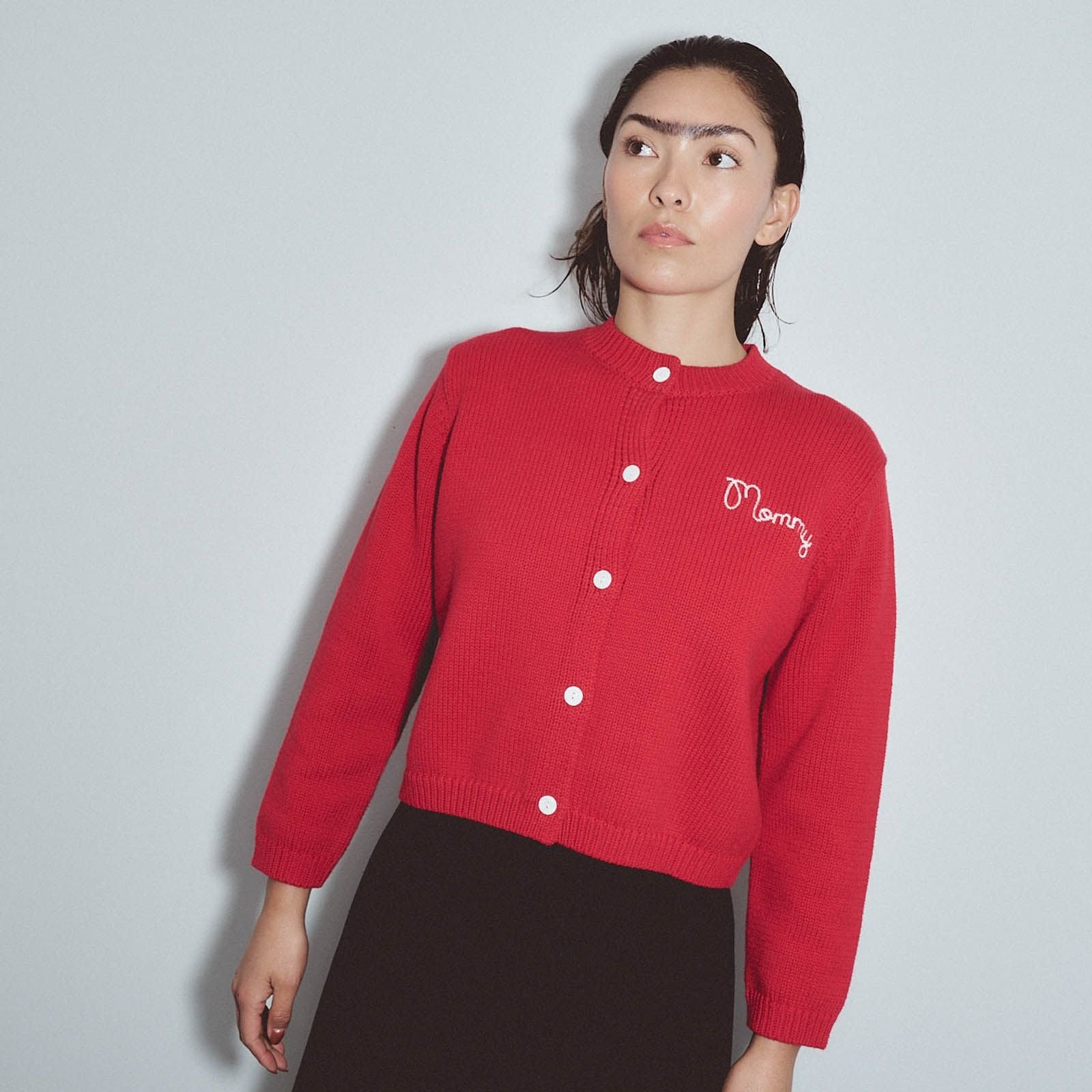 A woman wearing a red ribbed knit cardigan with white buttons and a white embroidered 'Merry' logo on the chest, paired with a dark brown skirt, against a light gray background. Mr. Rogers Mommy Cardigan by Fashion Brand Company.