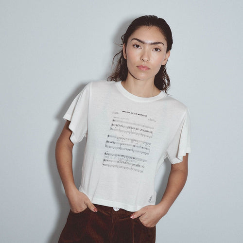 A woman wearing a white short-sleeve t-shirt with black musical notation text printed on the front, by Fashion Brand Company paired with dark brown corduroy pants, standing against a light gray background.
