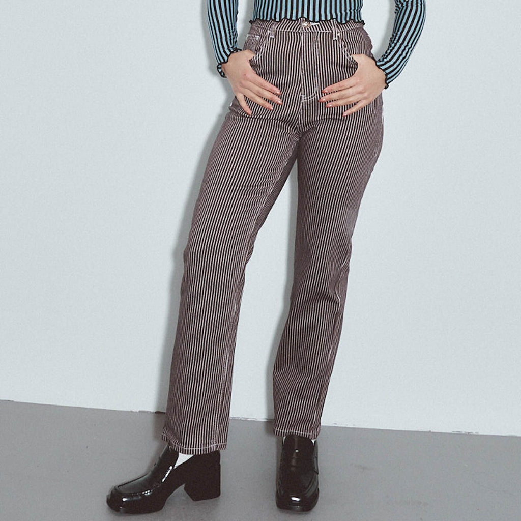 A model wearing brown and white vertically striped high-waisted straight-leg jeans, paired with a blue and white striped long-sleeve top and black patent leather boots, standing against a light gray wall. Thin Railroad stripe jeans by Fashion Brand Company.
