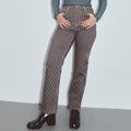 A model wearing brown and white vertically striped high-waisted straight-leg jeans, paired with a blue and white striped long-sleeve top and black patent leather boots, standing against a light gray wall. Thin Railroad stripe jeans by Fashion Brand Company.