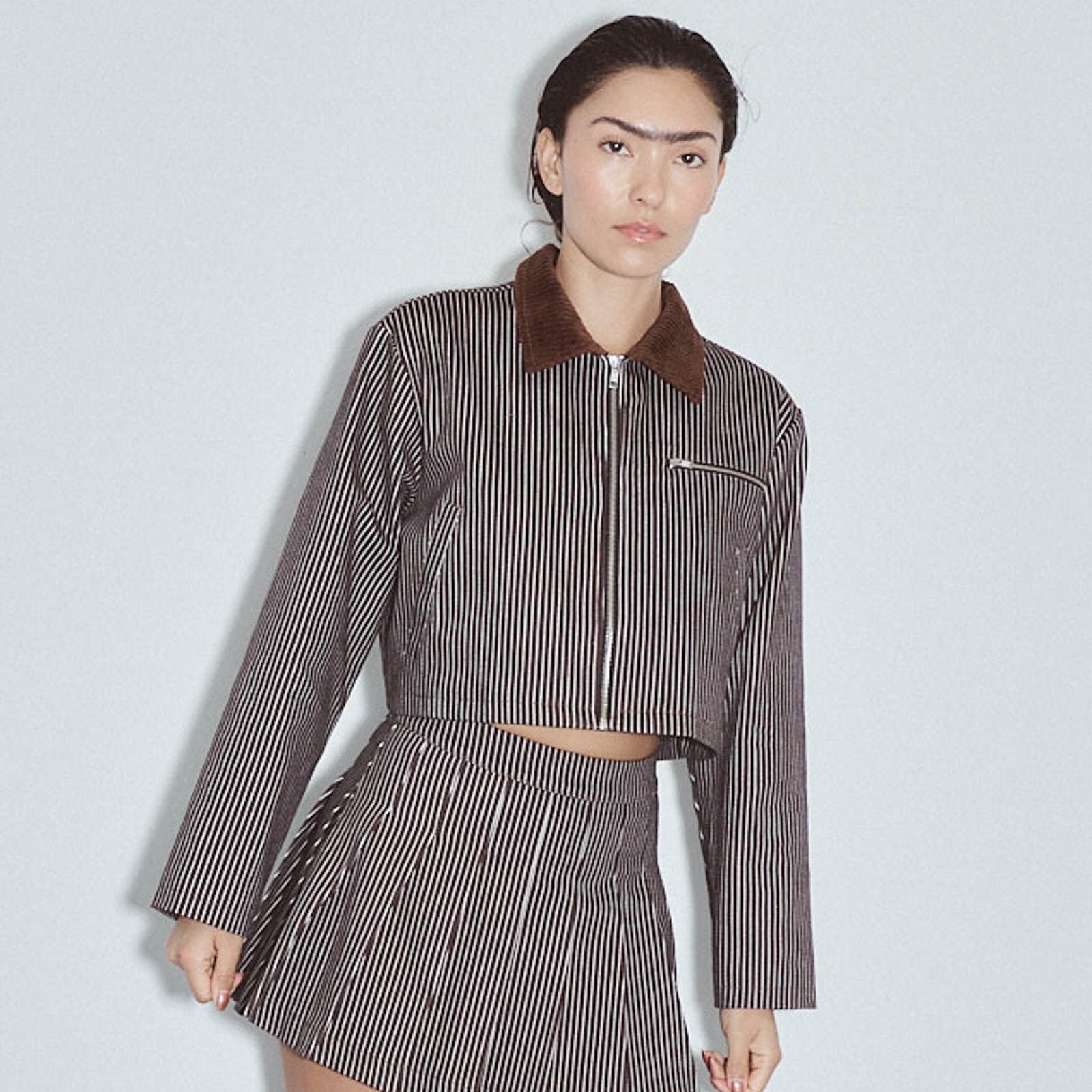 A model wearing a brown and white vertically striped cropped jacket with a brown corduroy collar and a matching striped mini skirt, set against a light gray studio background. Denim Construction Crop Jacket Railroad Stripe by Fashion Brand Company.