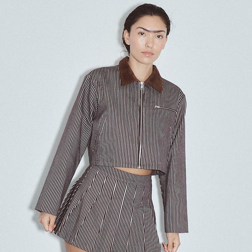 A model wearing a brown and white vertically striped cropped jacket with a brown corduroy collar and a matching striped mini skirt, set against a light gray studio background. Denim Construction Crop Jacket Railroad Stripe by Fashion Brand Company.