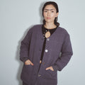 A woman wearing a dark purple windowpane plaid quilted jacket over a black top with a cutout, standing against a light gray wall. Survival Linen Quilted Jacket by Fashion Brand Company.