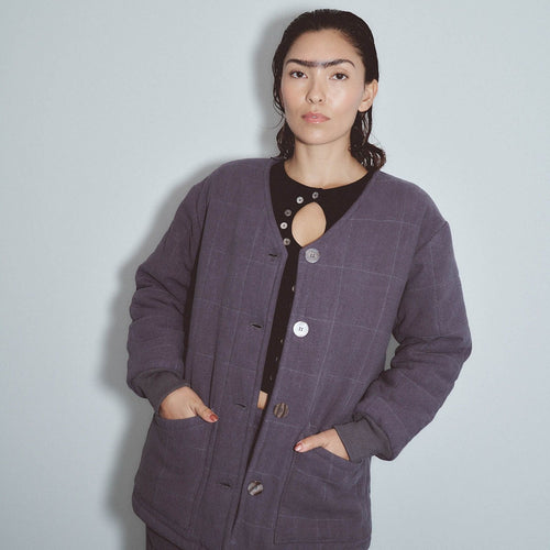 A woman wearing a dark purple windowpane plaid quilted jacket over a black top with a cutout, standing against a light gray wall. Survival Linen Quilted Jacket by Fashion Brand Company.