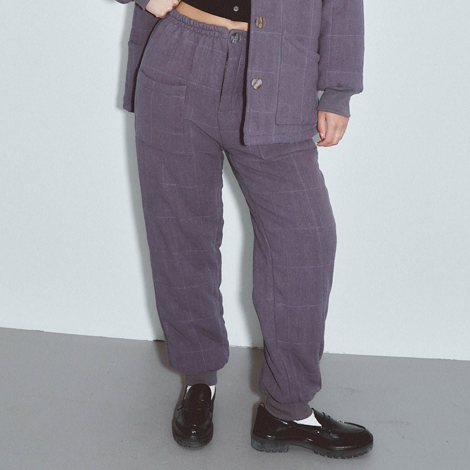 A person wearing a matching two-piece outfit consisting of a cropped button-front jacket and wide-leg corduroy pants in a muted purple/plum color, paired with black loafers against a plain light gray wall and floor. Survival Linen Quilted Pants by Fashion Brand Company.