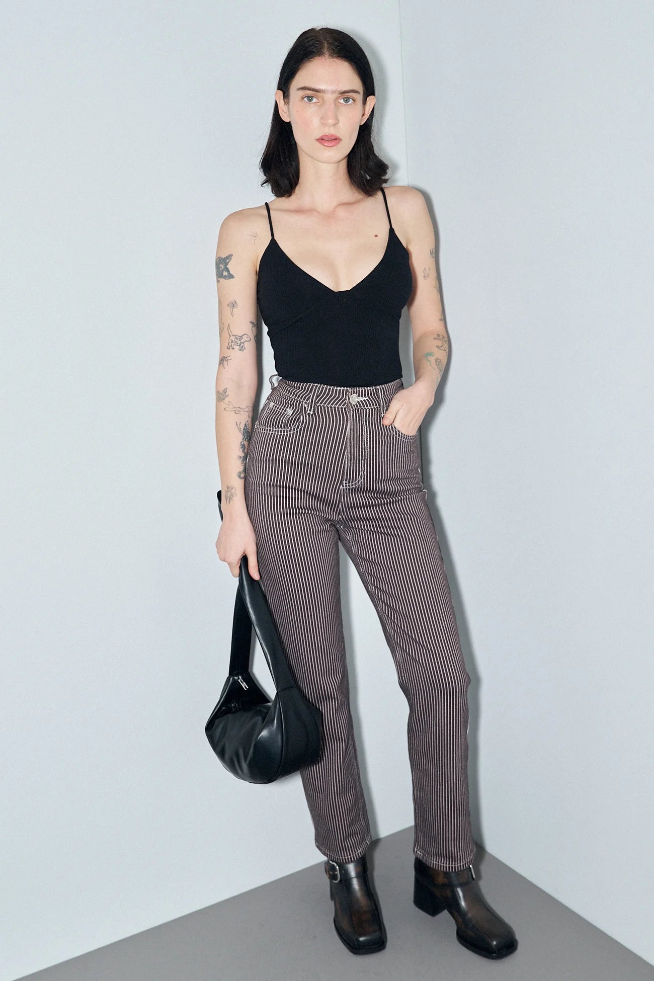 A model stands in a minimalist studio corner wearing a black spaghetti-strap camisole, brown pinstriped jeans, black ankle boots, and holding a black leather shoulder bag. Thin Railroad stripe jeans by Fashion Brand Company.