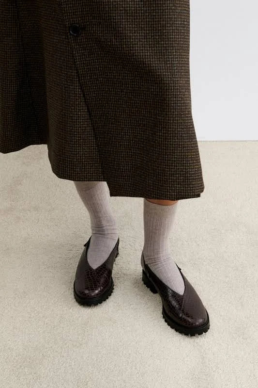 A woman wearing a pair of dark brown clog-style slingback Lenox Lug shoes by Rachel Comey with an embossed crocodile pattern, black platform soles, and adjustable ankle straps, shown against a light gray background.