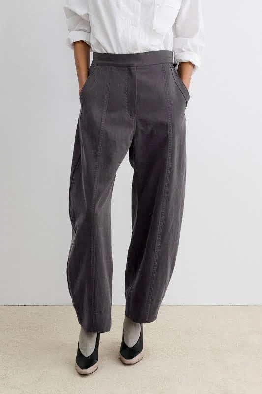 A person wearing dark charcoal gray barrel-leg midnight pants by Rachel Comey, styled with a white button-down shirt and black slip-on shoes, standing against a light gray wall and beige floor.