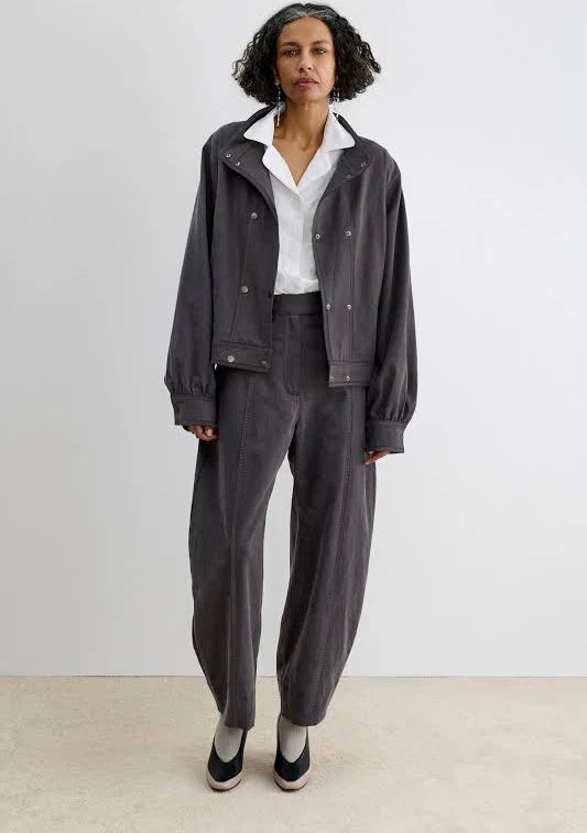 A model stands against a plain light gray studio wall wearing a dark charcoal gray corduroy jacket and matching barrel-leg Midnight Pant by Rachel Comey over a white collared shirt.