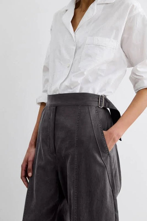 A close-up of a person wearing dark charcoal gray barrel-leg midnight pant by Rachel Comey and white button-up shirt, shown against a plain light gray background.