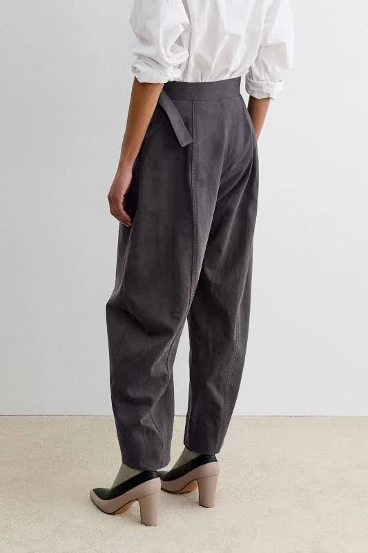 A person shown from the back wearing dark charcoal gray barrel-leg midnight pants by Rachel Comey, a white long-sleeve blouse, and black-and-beige heels against a plain light wall and beige floor.