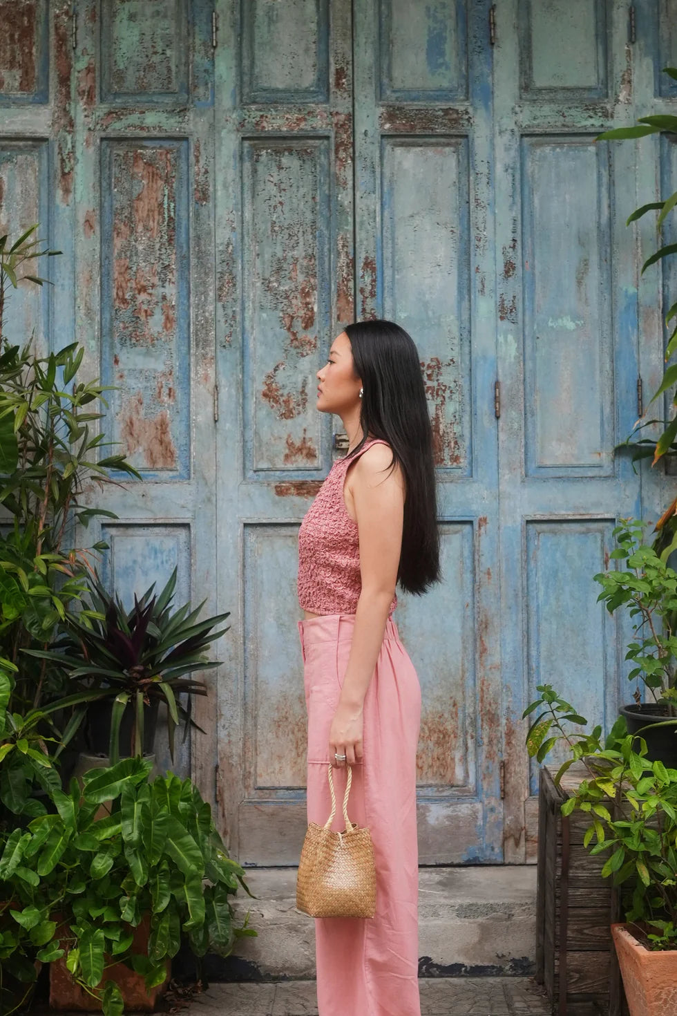 Woman in pink outfit standing in front of a weathered wooden door with plants around.