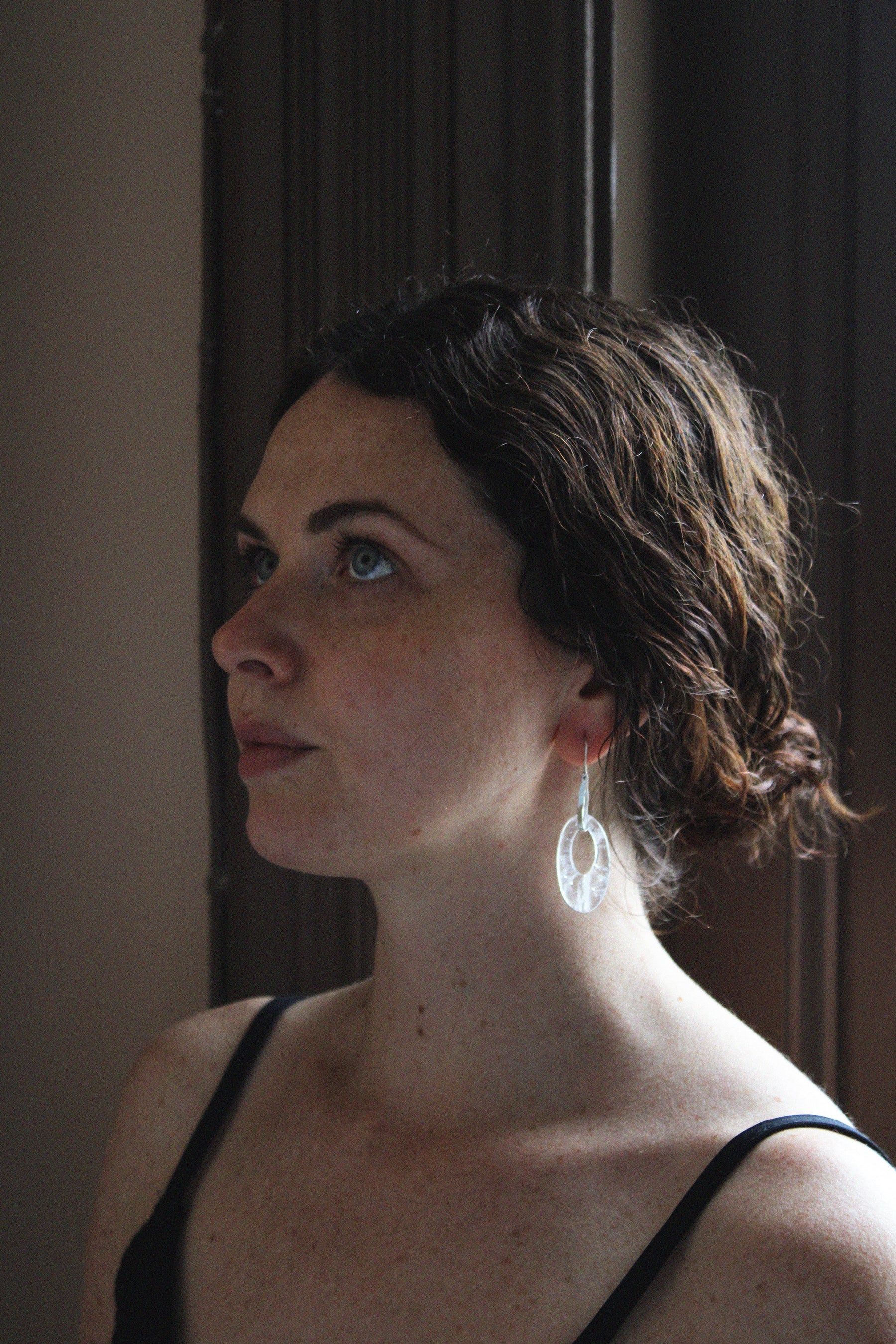 A woman in profile wearing a clear, teardrop-shaped dangle moonlight earring by Takara, photographed in warm, dim indoor light.