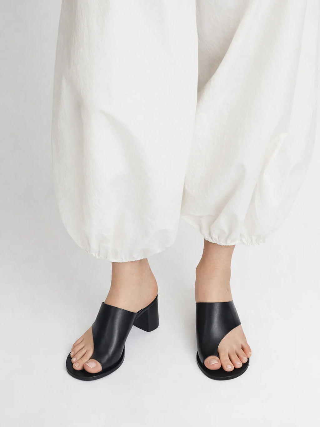 Woman wearing black cutout Rosate sandal by ATP Atelier with a high heel with white pants on a white background