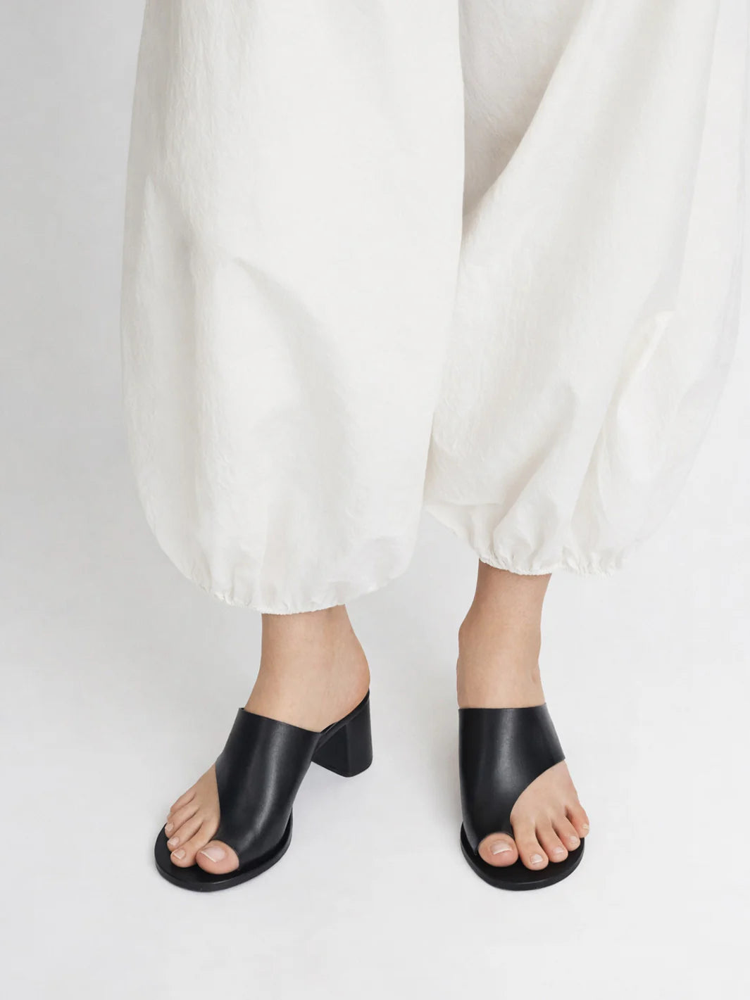 Woman wearing black cutout Rosate sandal by ATP Atelier with a high heel with white pants on a white background