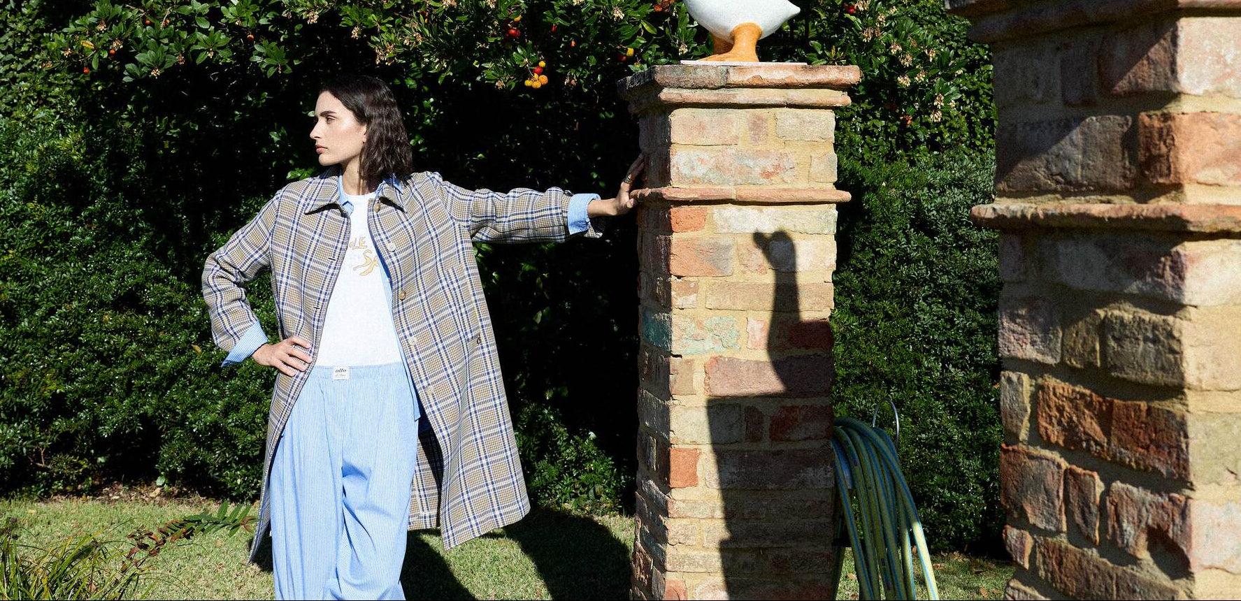 Woman in a plaid coat and blue pants standing next to a brick column with a garden background