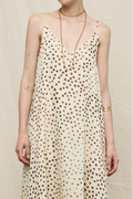 A woman wearing a cream-colored spaghetti-strap dress with a brown polka dot pattern, styled with gold jewelry against a neutral beige background.