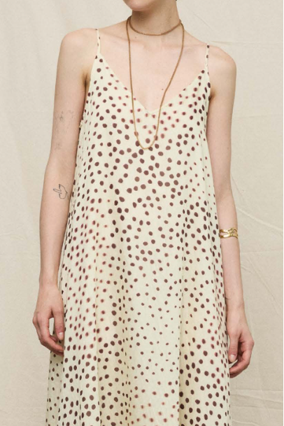 A woman wearing a cream-colored spaghetti-strap dress with a brown polka dot pattern, styled with gold jewelry against a neutral beige background.