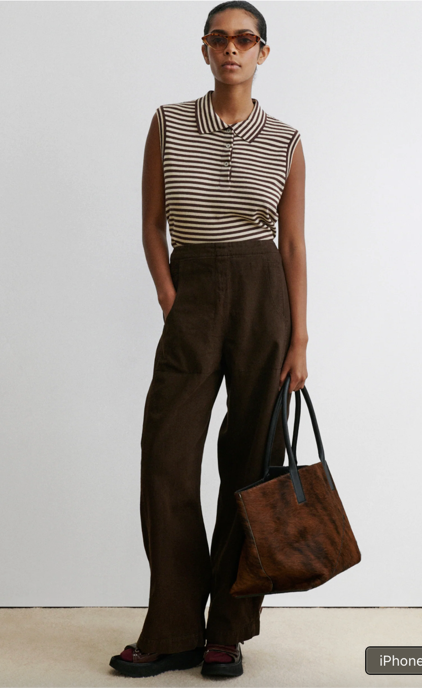 Person wearing a striped sleeveless top and brown pants, holding a brown bag against a white background.