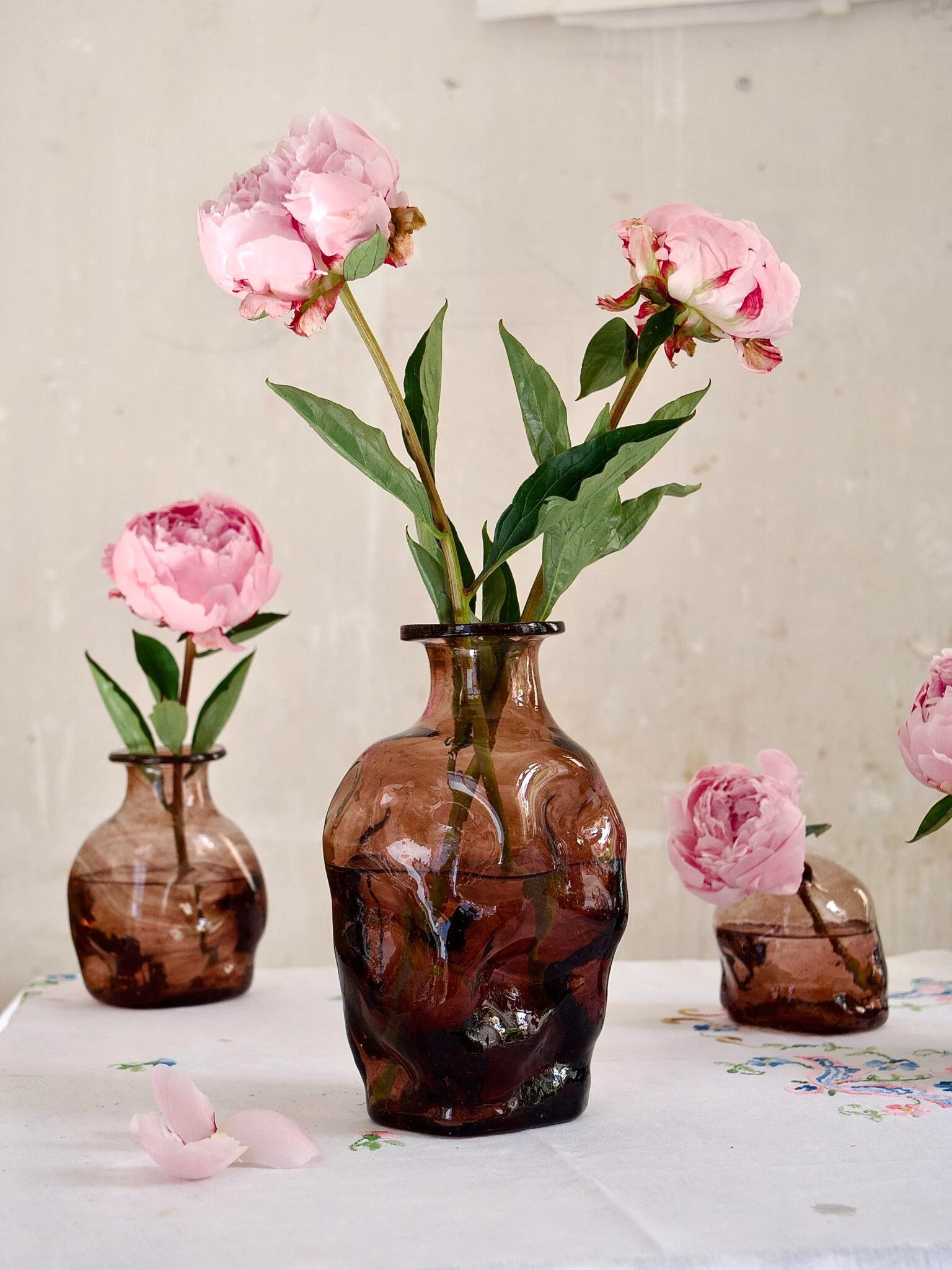 Three brown glass vases with pink flowers on a light surface.