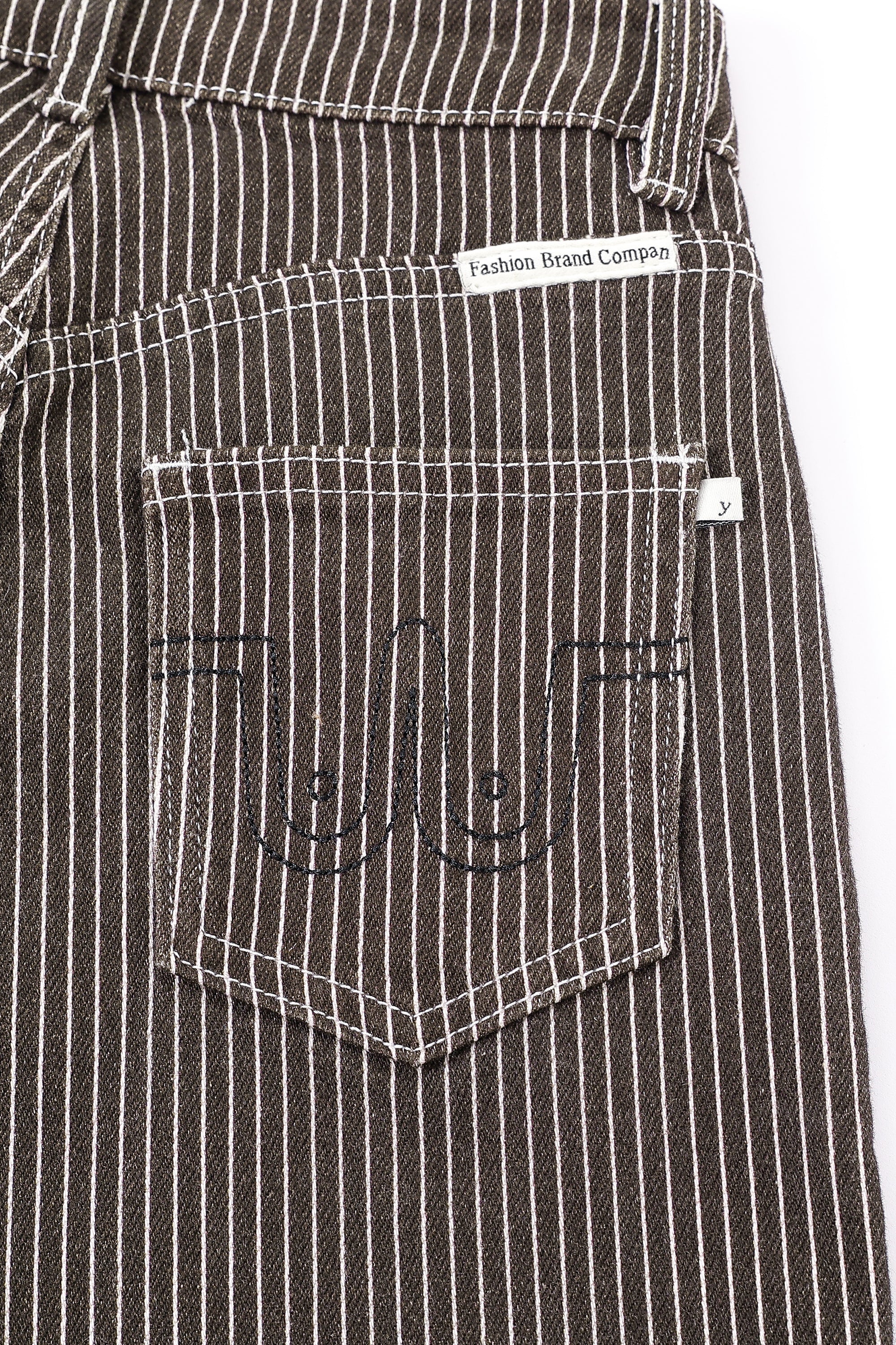 Close-up of the back pocket of dark brown pinstriped jeans with white vertical stripes, featuring black embroidered stylized boobs and a white brand label. Thin Railroad stripe jeans by Fashion Brand Company.