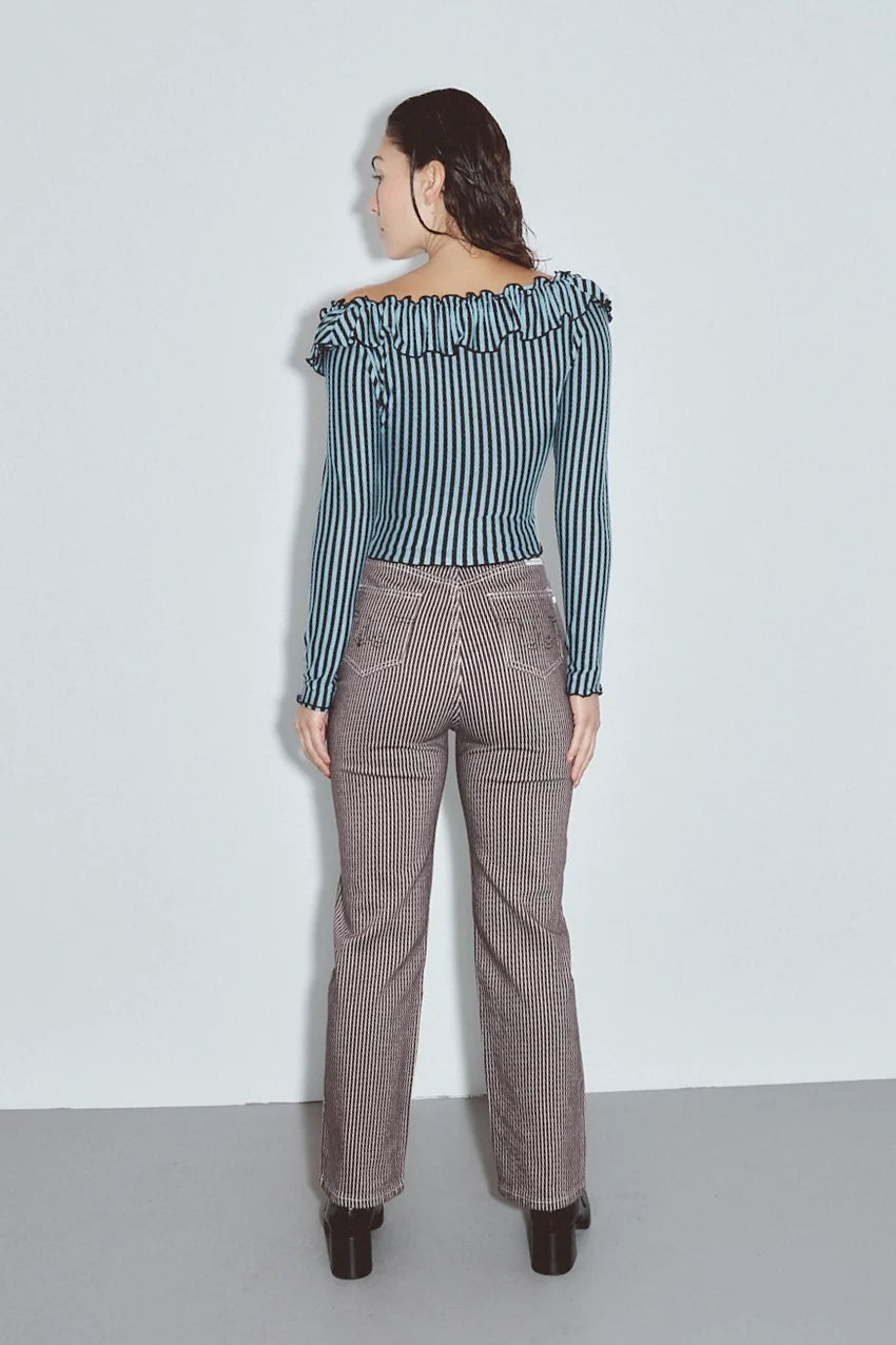 A woman is shown from the back wearing a blue and white vertically striped off-the-shoulder long-sleeve top and brown pinstriped trousers, standing against a plain light gray wall. Thin Railroad stripe jeans by Fashion Brand Company.