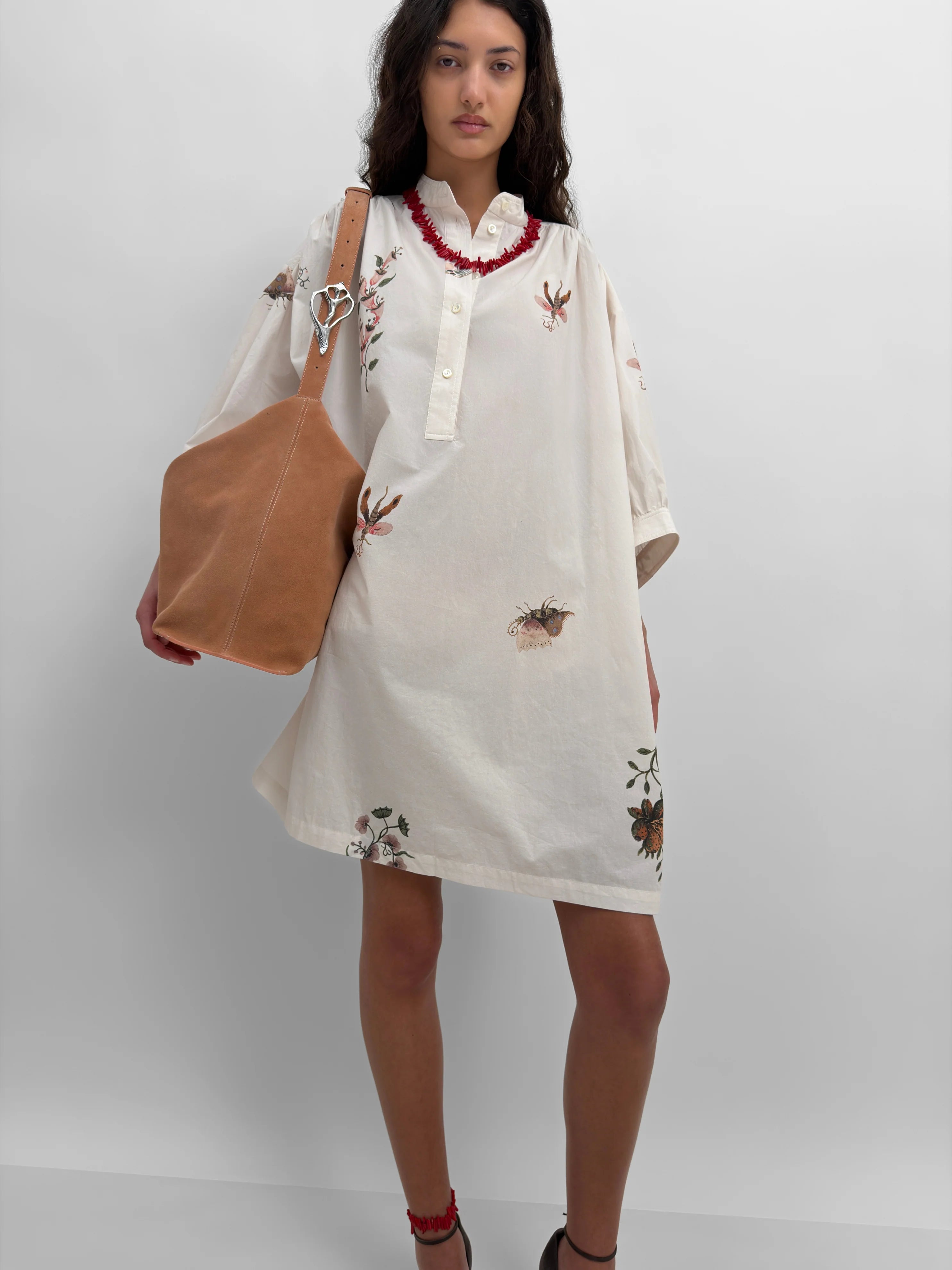 Woman wearing a white printed dress with floral patterns holding a brown bag against a white background